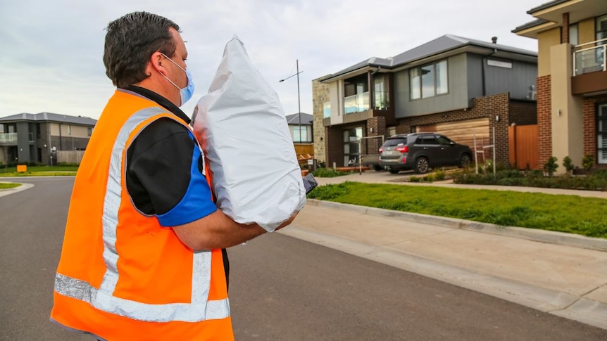 Man wearing high vis jacket walks towards a house carrying a white parcel.