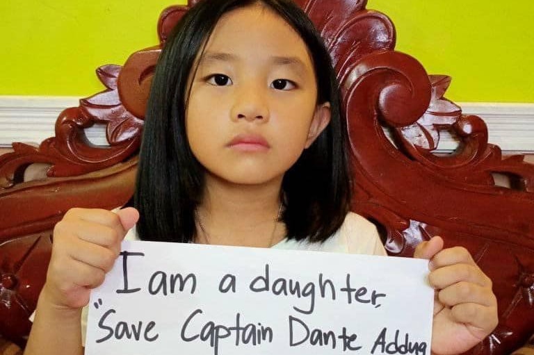 A young Filipino girl sitting on a wooden chair holding a sign saying: 'I am a daughter, Save Captain Dante Addng and his crew'