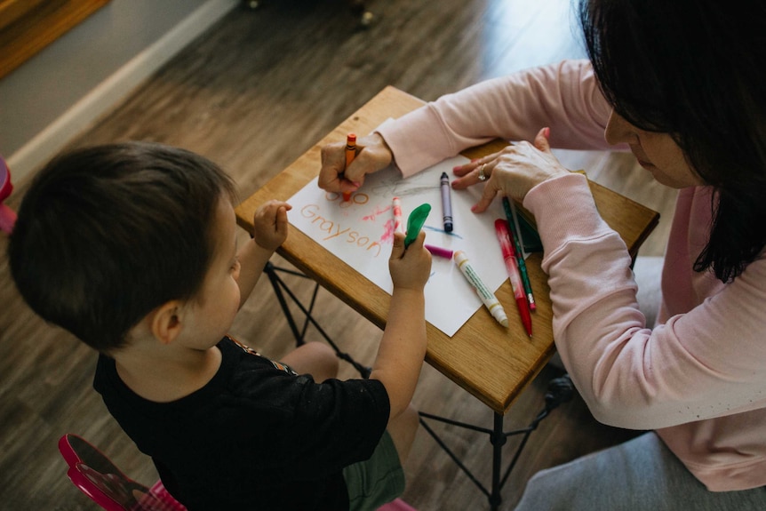 Toddler sitting at small table and doing some drawing alongside an adult