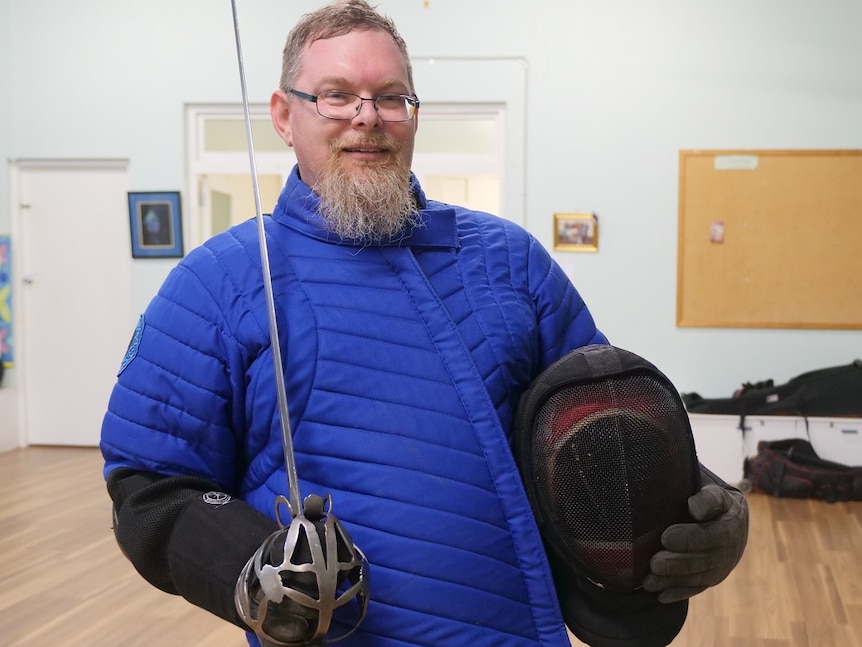 Man with bushy red beard holds a helmet in one hand and sword in the other
