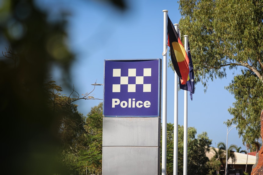 A police sign surrounded by trees, with a row of flags behind it
