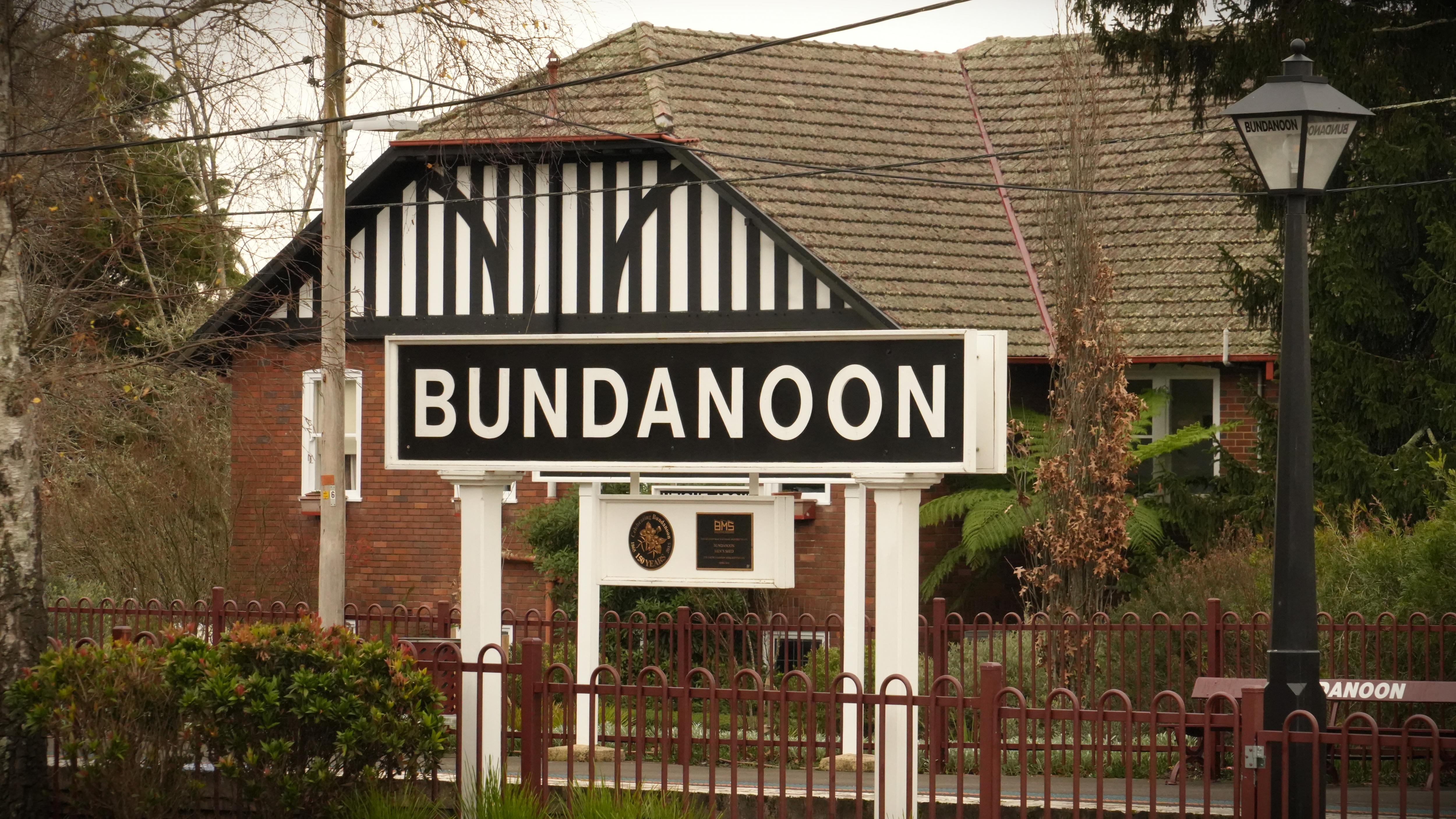 A train station sign reading "Bundanoon" in the foreground, and a Tudor-looking property in the background.