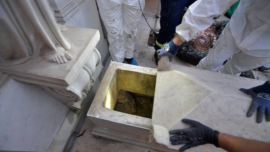 Looking down on a marble tomb, workers in hazmat suits and blue gloves pull its top back to reveal no remains left inside.