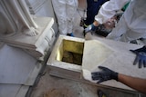 Looking down on a marble tomb, workers in hazmat suits and blue gloves pull its top back to reveal no remains left inside.