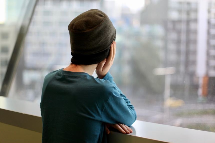 An unidentifiable teenage boy, wearing a beanie, stares out a window.