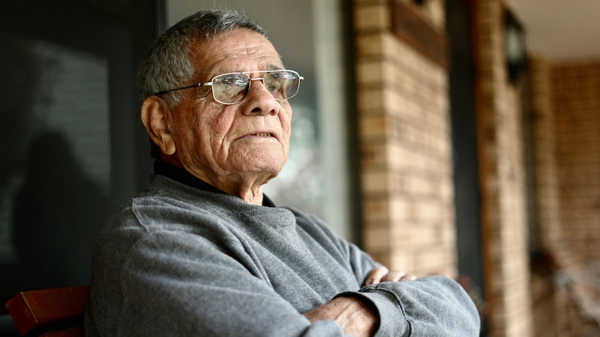 Older man with grey hair and glasses sits on his porch in a grey jumper with his arms crossed