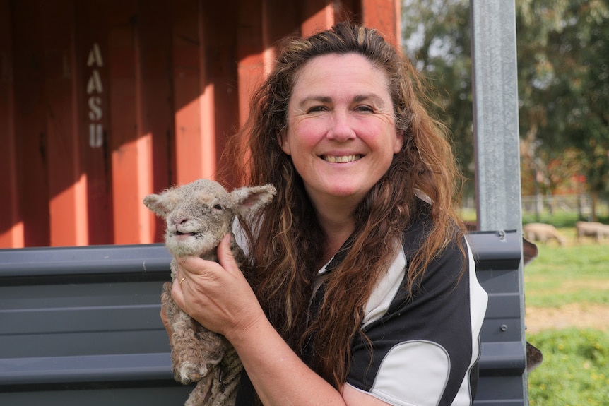 A woman with brown hair holds a small lamb in her hands and smiles at the camera.