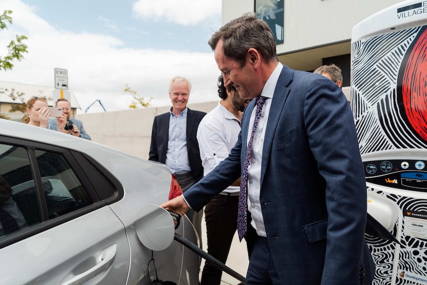 Premier Mark McGowan plugs in an electric vehicle at a charging station with several people in the background.