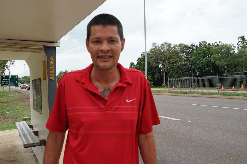 A man in a red shirt smiles at the camera in front of the Stuart Highway next to his flat.