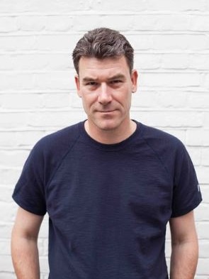 Man in a blue-t-shirt stands in front of a white brick wall.