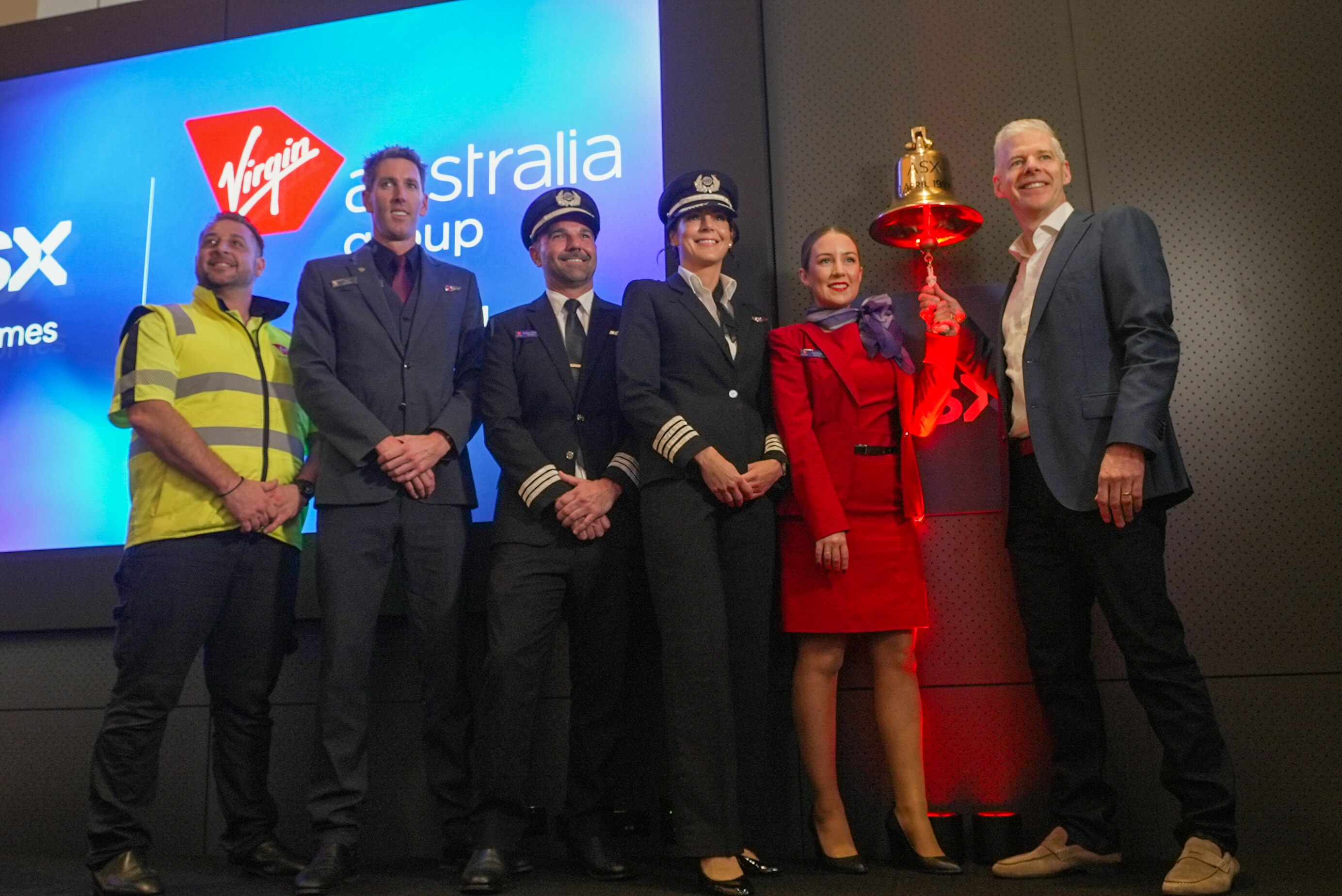 Five people pose with a gold bell, including a grounds worker wearing fluro, two pilots and a flight attendant.