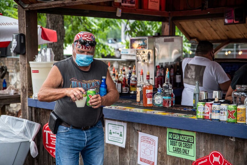 A man in a face mask and US flag bandana on his head carries two drinks away from an al fresco bar in Texas