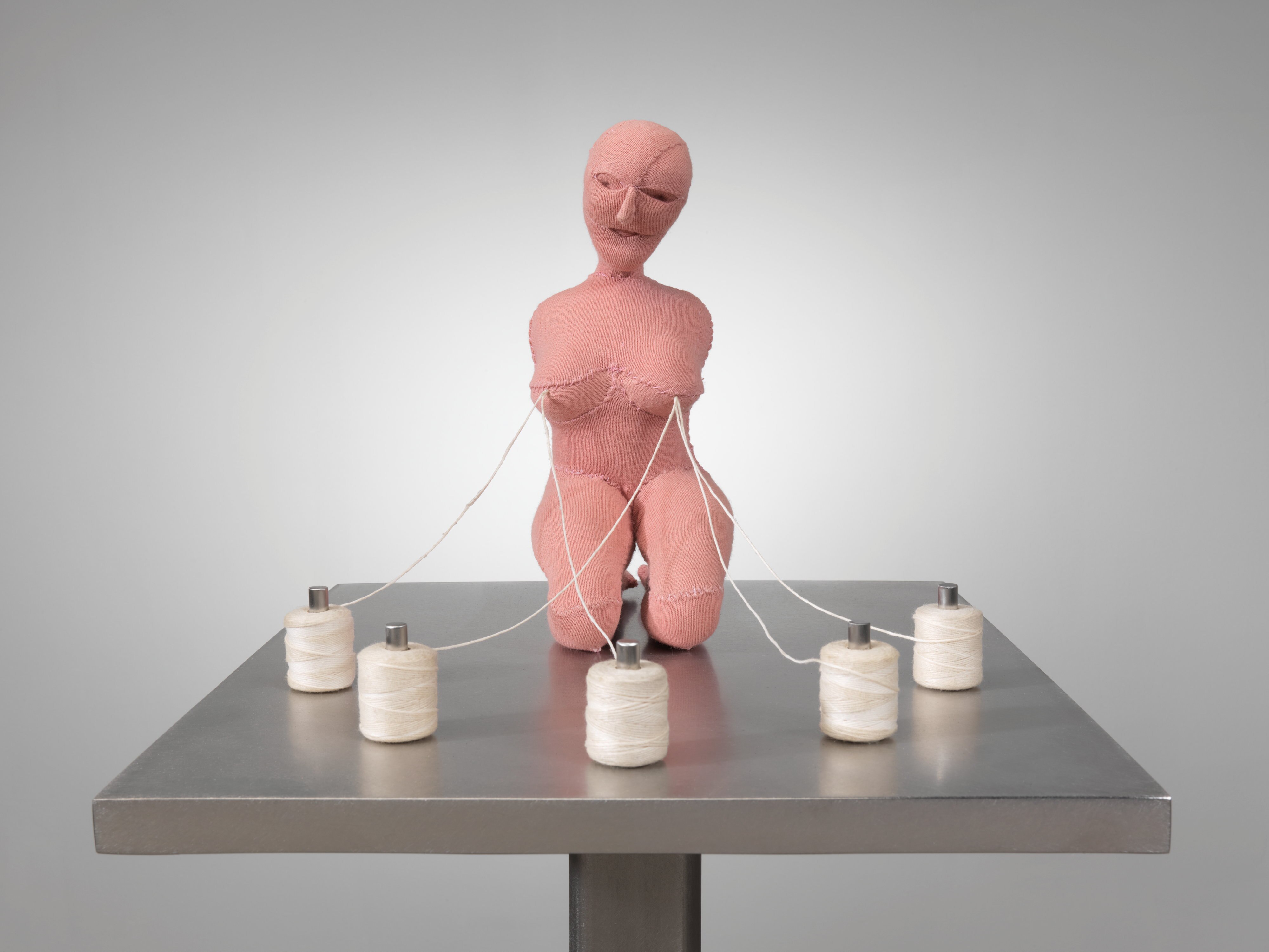 A fabric statue of a naked woman surrounded by five white sewing thread spools with thread spilling from her breasts, like milk