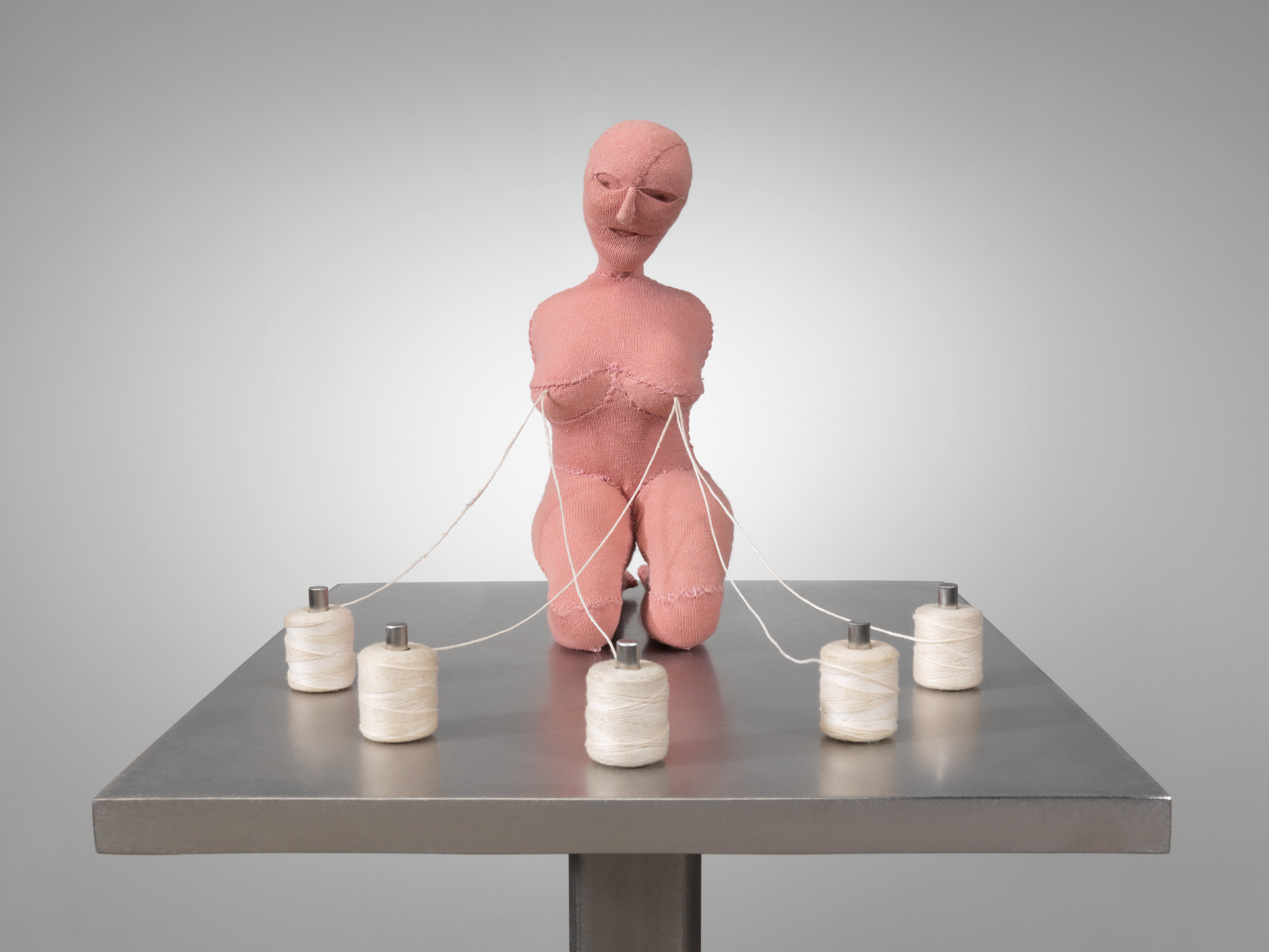 A fabric statue of a naked woman surrounded by five white sewing thread spools with thread spilling from her breasts, like milk