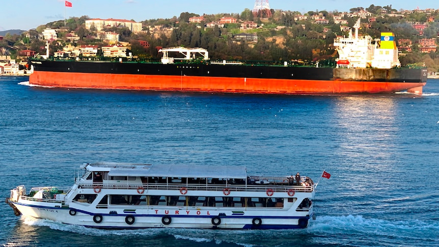 A black and orange oil tanker sails past a hilly city and a small white ferry in daylight.