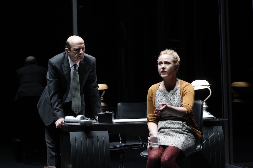 Balding middle-aged white man in suit leans over desk and looks at young white blonde woman in dress and tights holding water