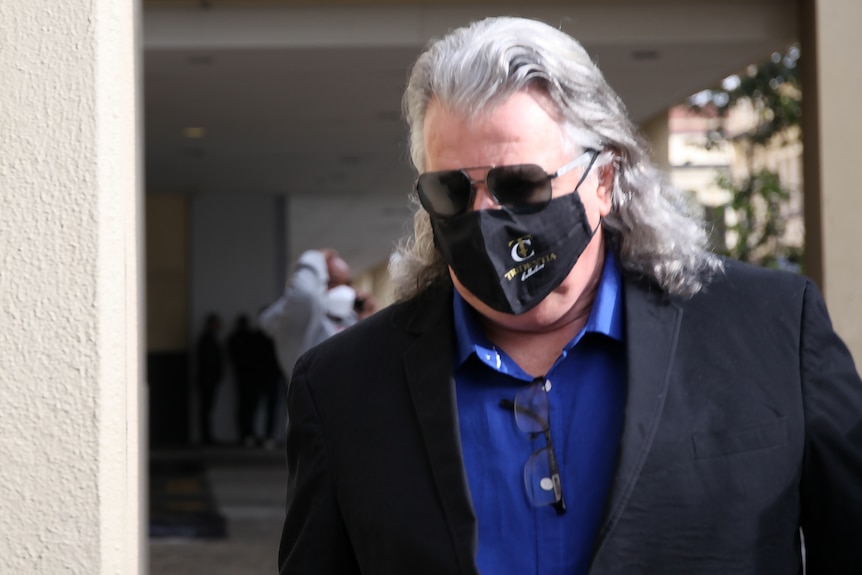 A tight mid shot of a man with long greying hair, sunglasses and a dark suit and shirt on walking outside court in Perth.