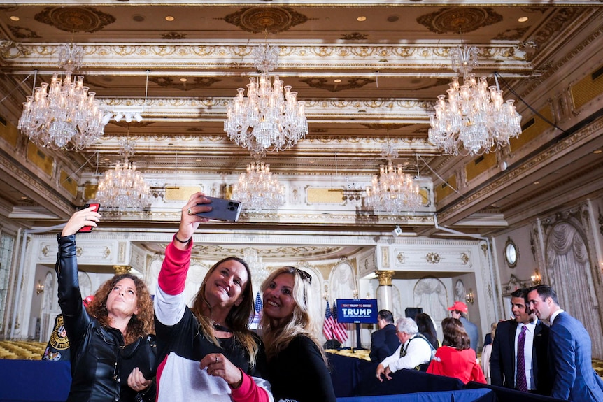 Three women hold their phones high to take a selfie inside an ornate ballroom
