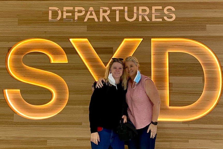 Interior scene Poppy Starr Olsen and mum Thomas in front of sign that says Syd Departures