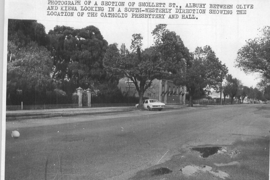 A black and white photo, with typed notes, showing a street in a country town. A white car is parked under a tree.