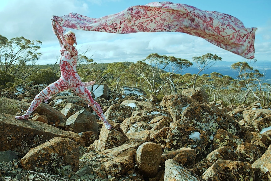 A woman in a red and white body suit stands on rocks holding a matching length of fabric blowing in the wind.