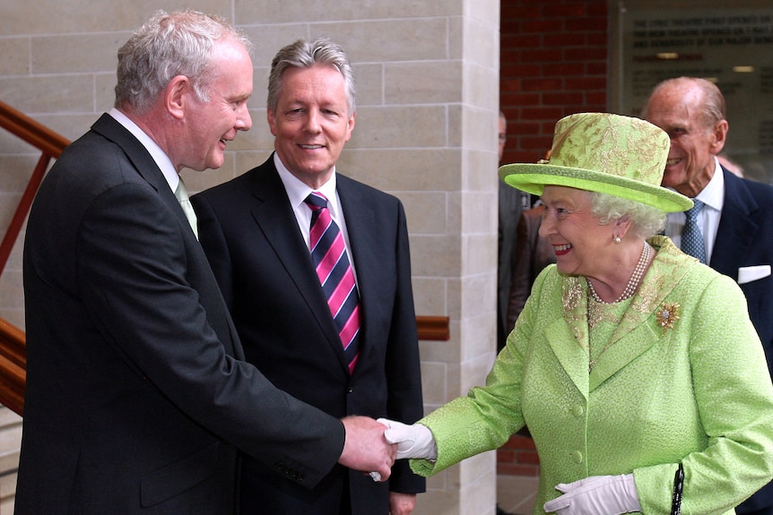 Britain's Queen Elizabeth II wears a green hat and matching twin set as two suited men watch her shake hands with a third man.