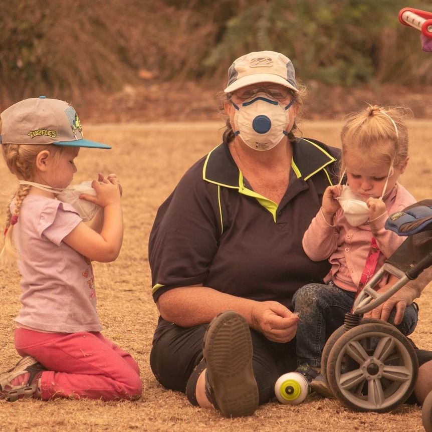 A woman with a face mask sits on the ground with two young children who are pulling off their masks. A trolley sits nearby.