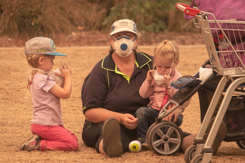 A woman with a face mask sits on the ground with two young children who are pulling off their masks. A trolley sits nearby.