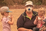 A woman with a face mask sits on the ground with two young children who are pulling off their masks. A trolley sits nearby.