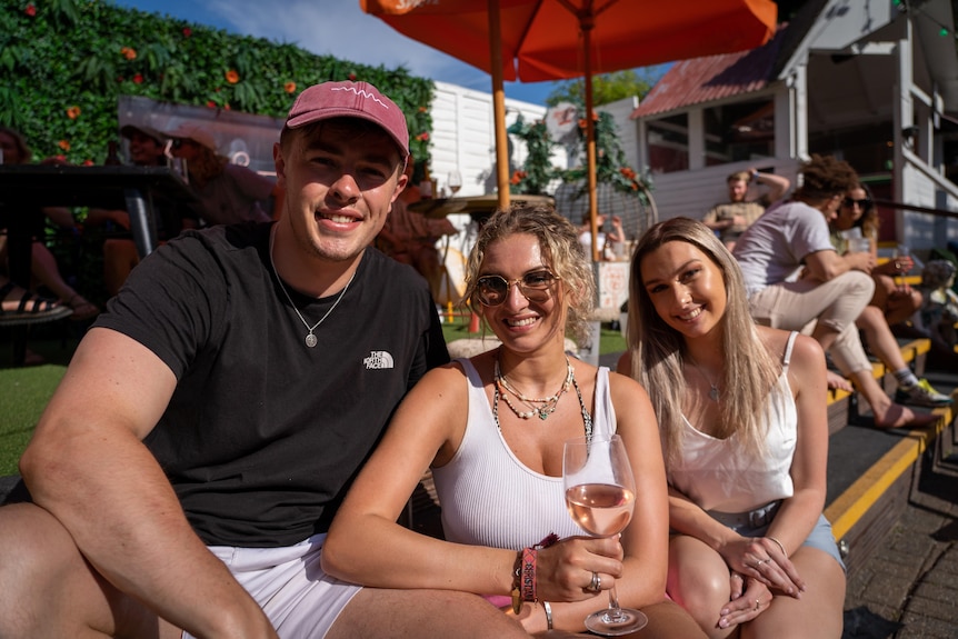 A young man in a hat and two young women pose for the camera in a beer garden.