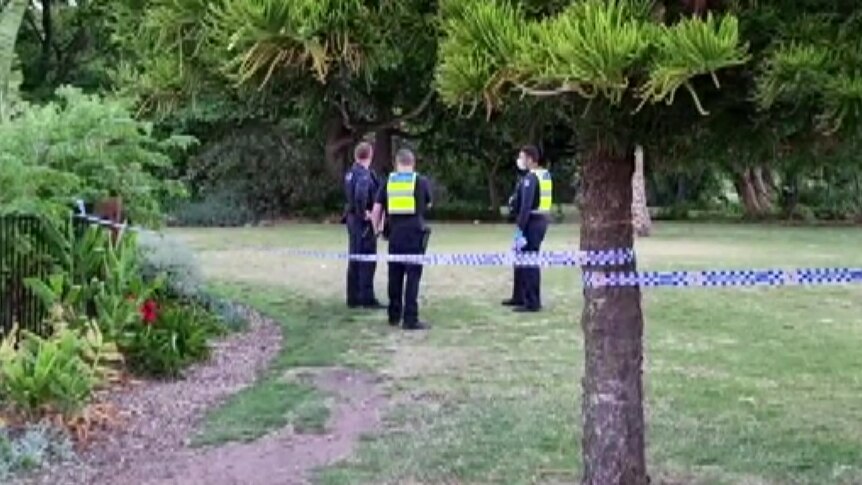 Police stand in Footscray Park, behind police tape wound around a tree.