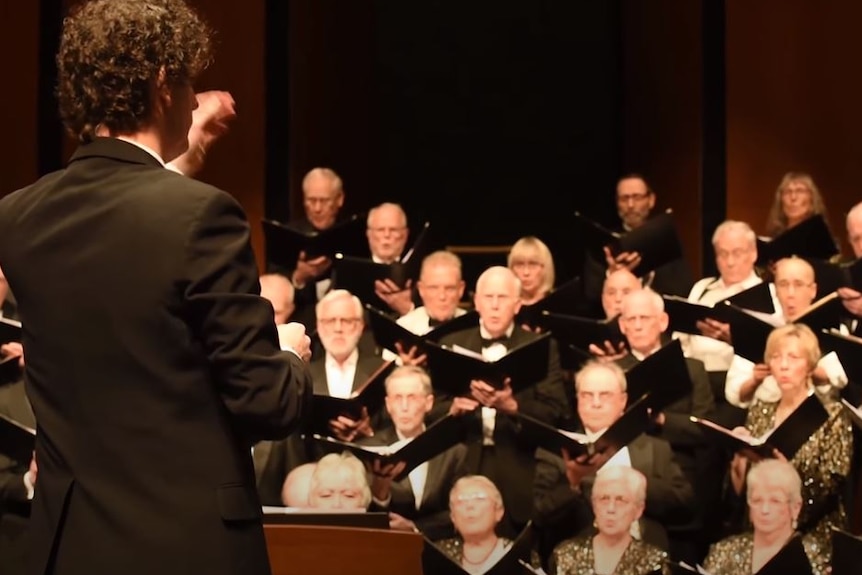 A conductor wearing a black suit standing in front of a choir.