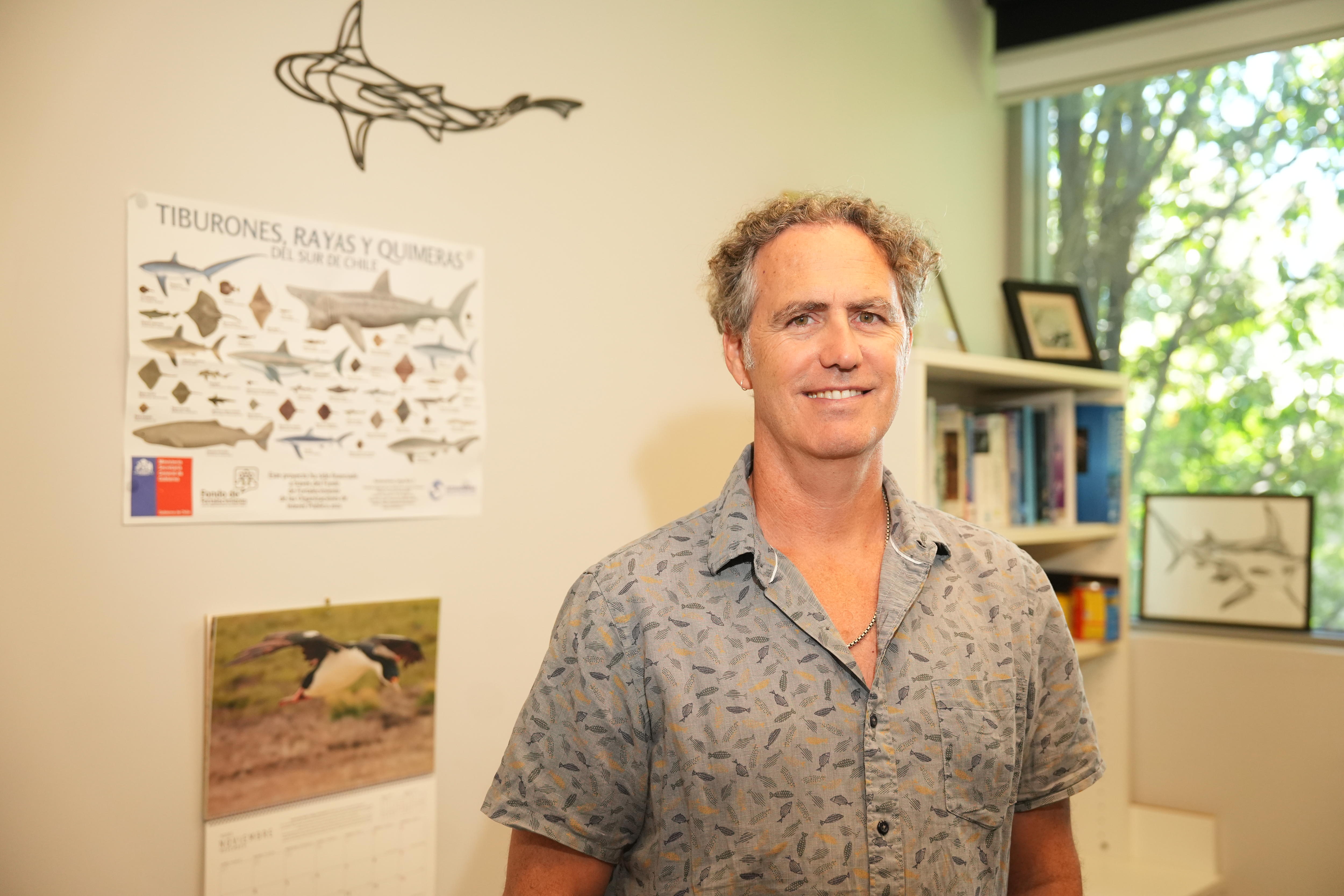 Un hombre con pantalones cortos de manga corta en una oficina con un cartel de tiburones en la pared.