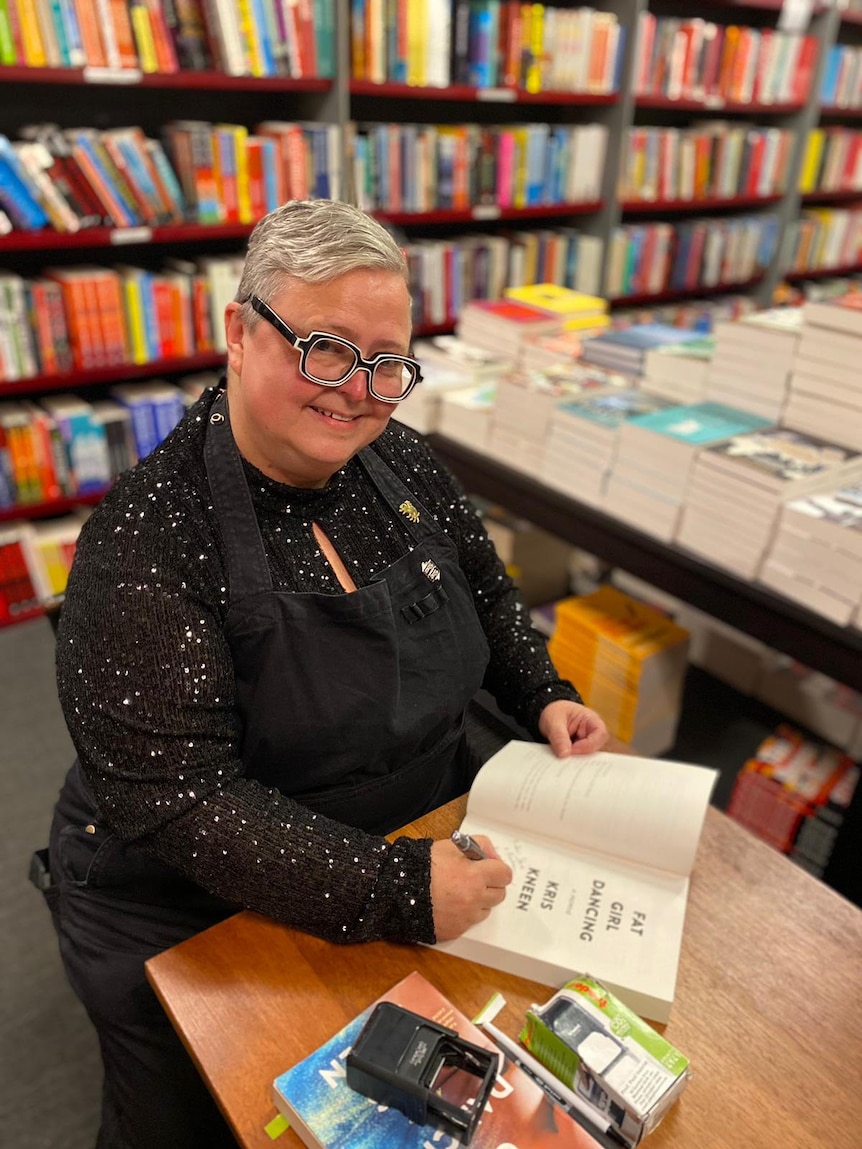 A person with short hair and thick glasses signs copies of a book in a bookstore.