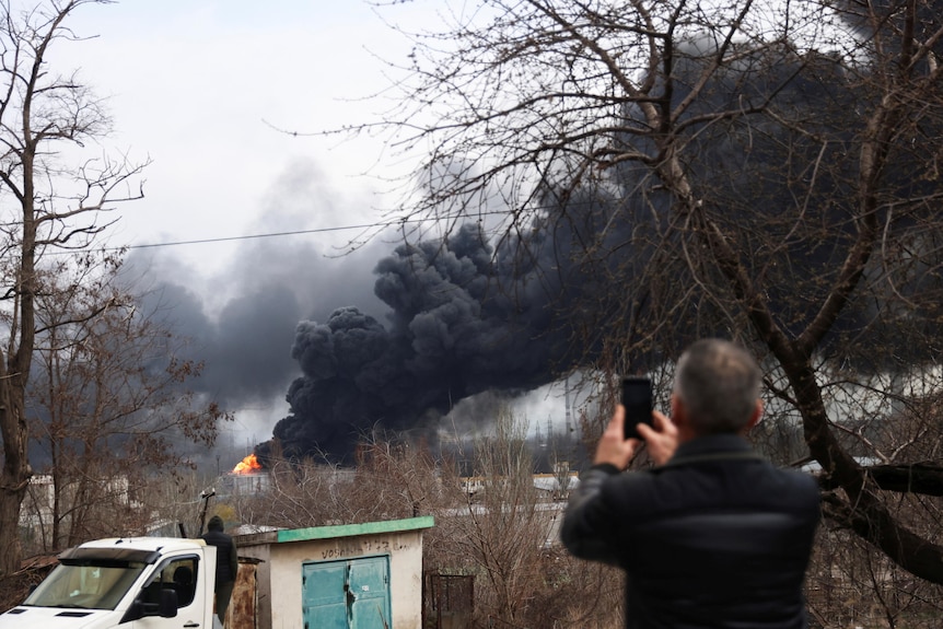 A man takes a photo of smoke from an oil refinery after a Russian missile attack.