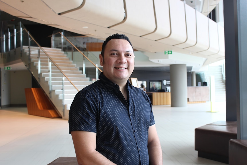 A man with short dark hair in a blue button-up shirt stands in a university foyer