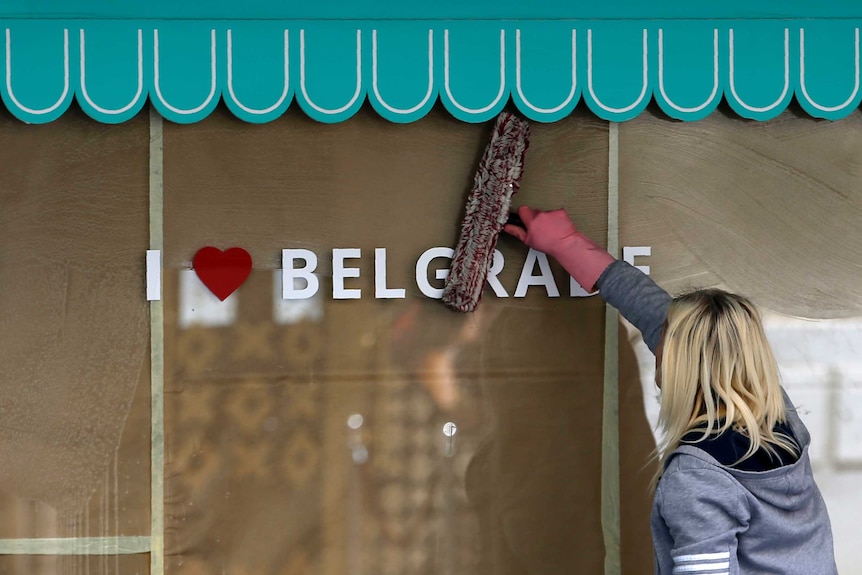 A woman drags a cleaning rag over a shop window. Decals on the window read 'I heart Belgrade'.