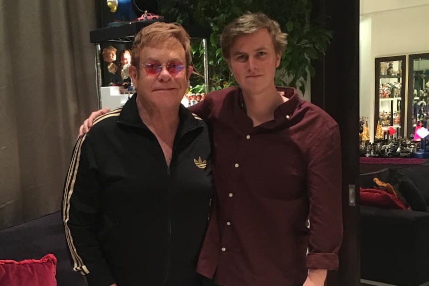 A man in a black Adidas tracksuit and pink glasses standing with a younger man in a red shirt.