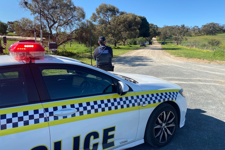 The back of a police officer leaning against a police car, parked in the middle of a dirt road