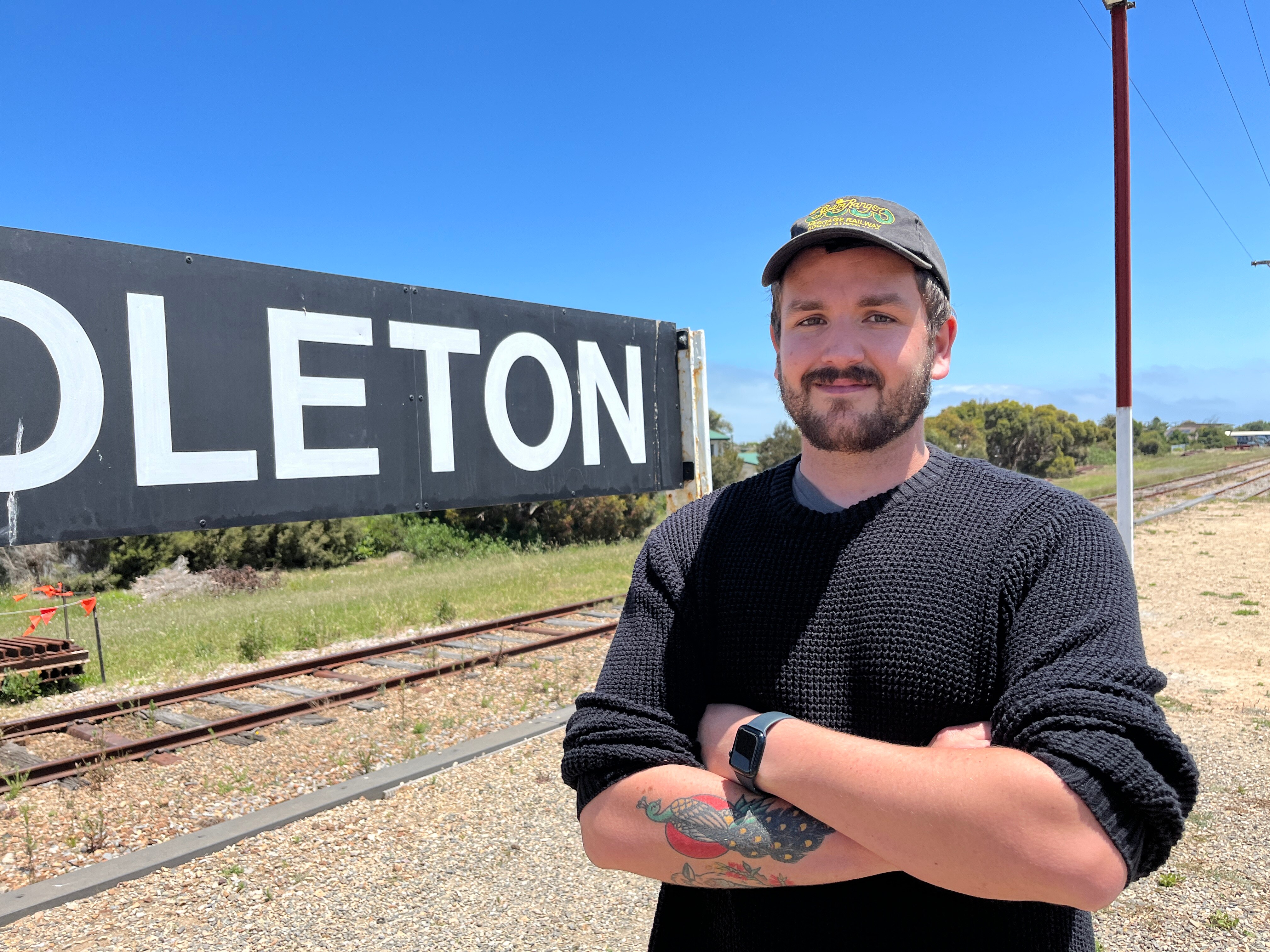 Man wearing dark shirt and cap stands with arms crossed in front of train line.