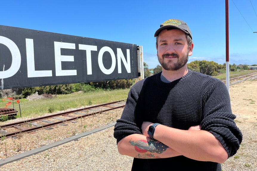 Man wearing dark shirt and cap stands with arms crossed in front of train line.