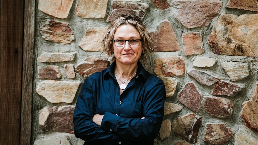 A white woman with glasses and wavy sandy-coloured hair wearing a navy shirt stands against a stone wall with arms crossed