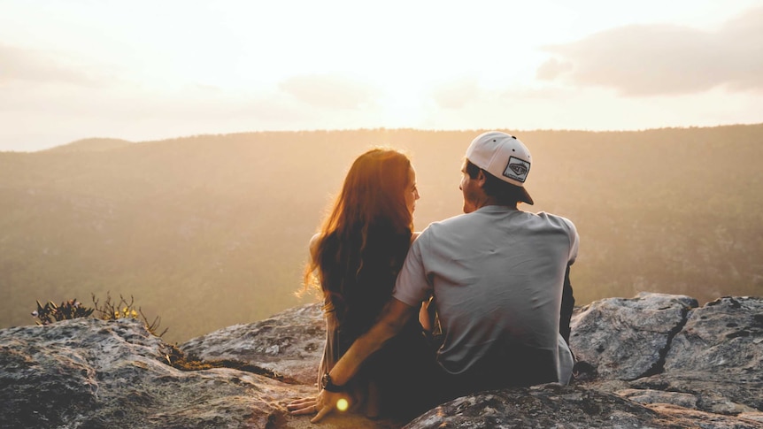 A man and woman sit on the edge of a cliff watching the sunset