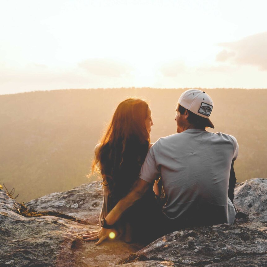 A man and woman sit on the edge of a cliff watching the sunset