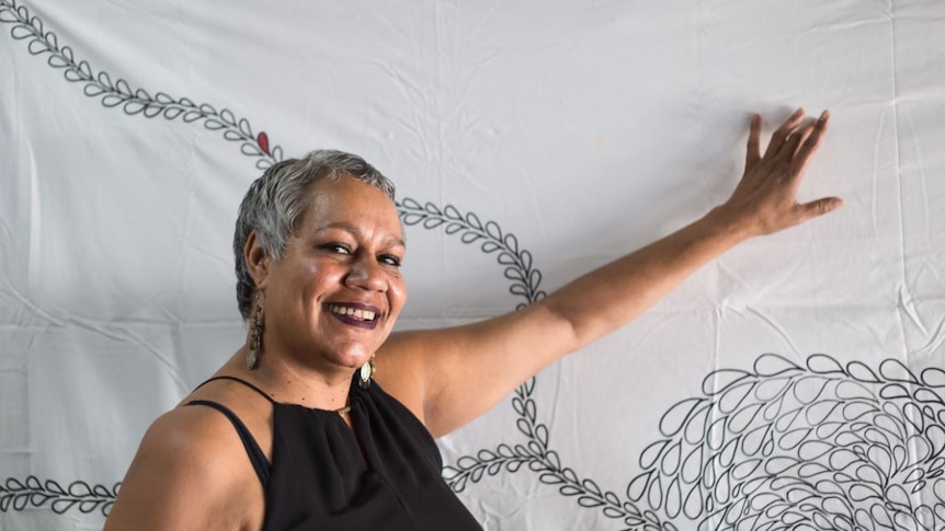Indigenous playwright Tessa Rose smiles in front of a white backdrop with a black line drawing