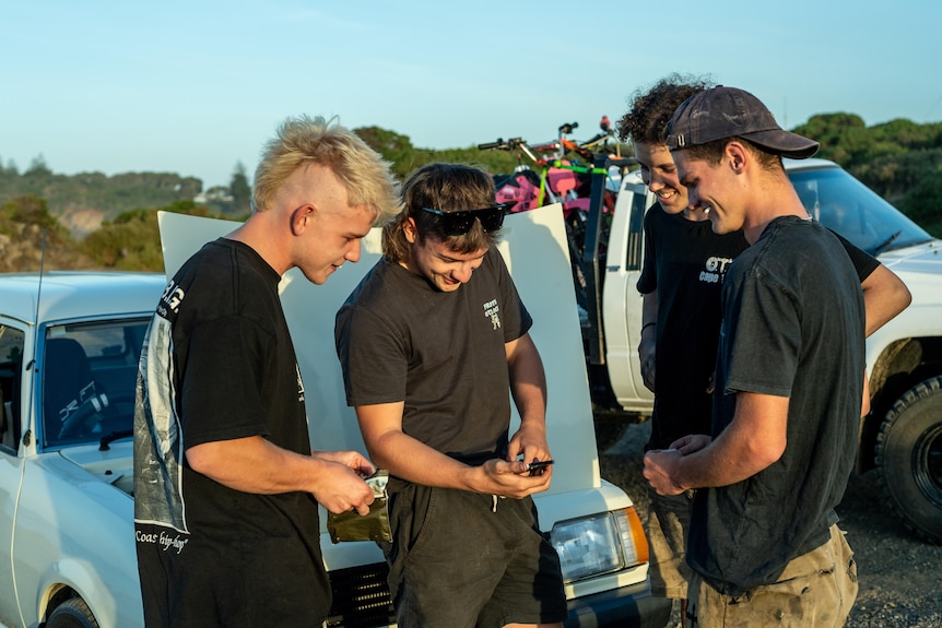 Boys gather around each other having a laugh next to their cars by the beach. 