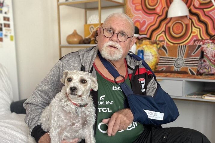 Robert Muir sits on his bed and holds his dog, his arm in a sling after shoulder surgery.