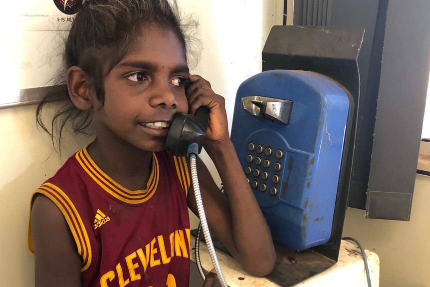 A young Aboriginal girl stands talking on a pay phone wearing a Cleveland Cavaliers LeBron Janes jersey.