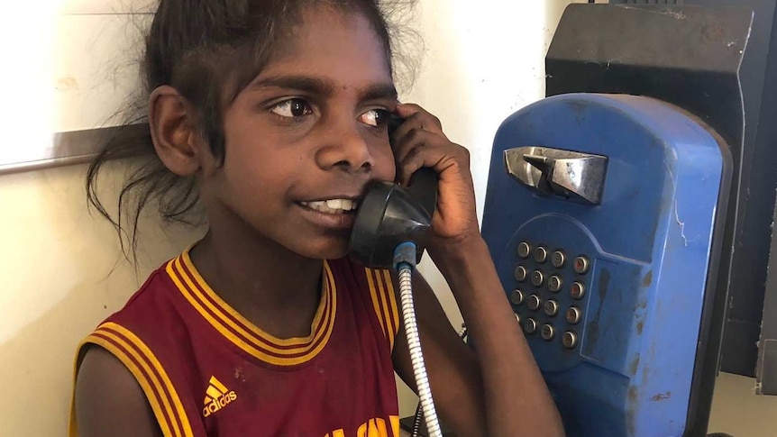 A young Aboriginal girl stands talking on a pay phone wearing a Cleveland Cavaliers LeBron Janes jersey.