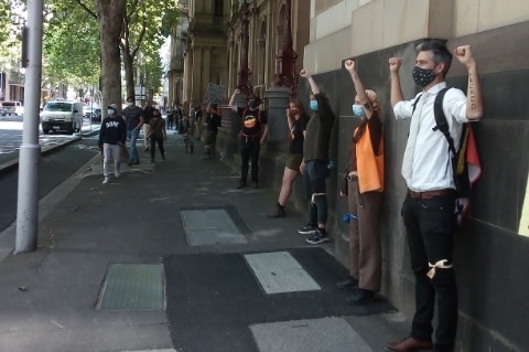 Masked people line up at regular intervals along with the wall of a state building with their fists in the air.